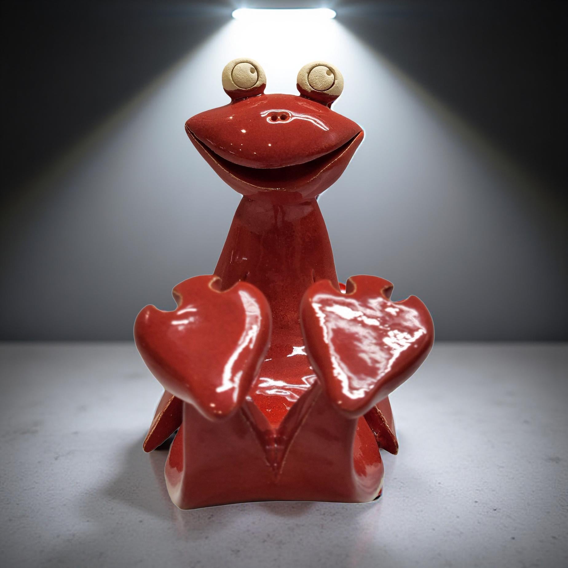 A red ceramic frog figurine with large eyes and a wide smile sits on a gray surface, illuminated by a bright light source.