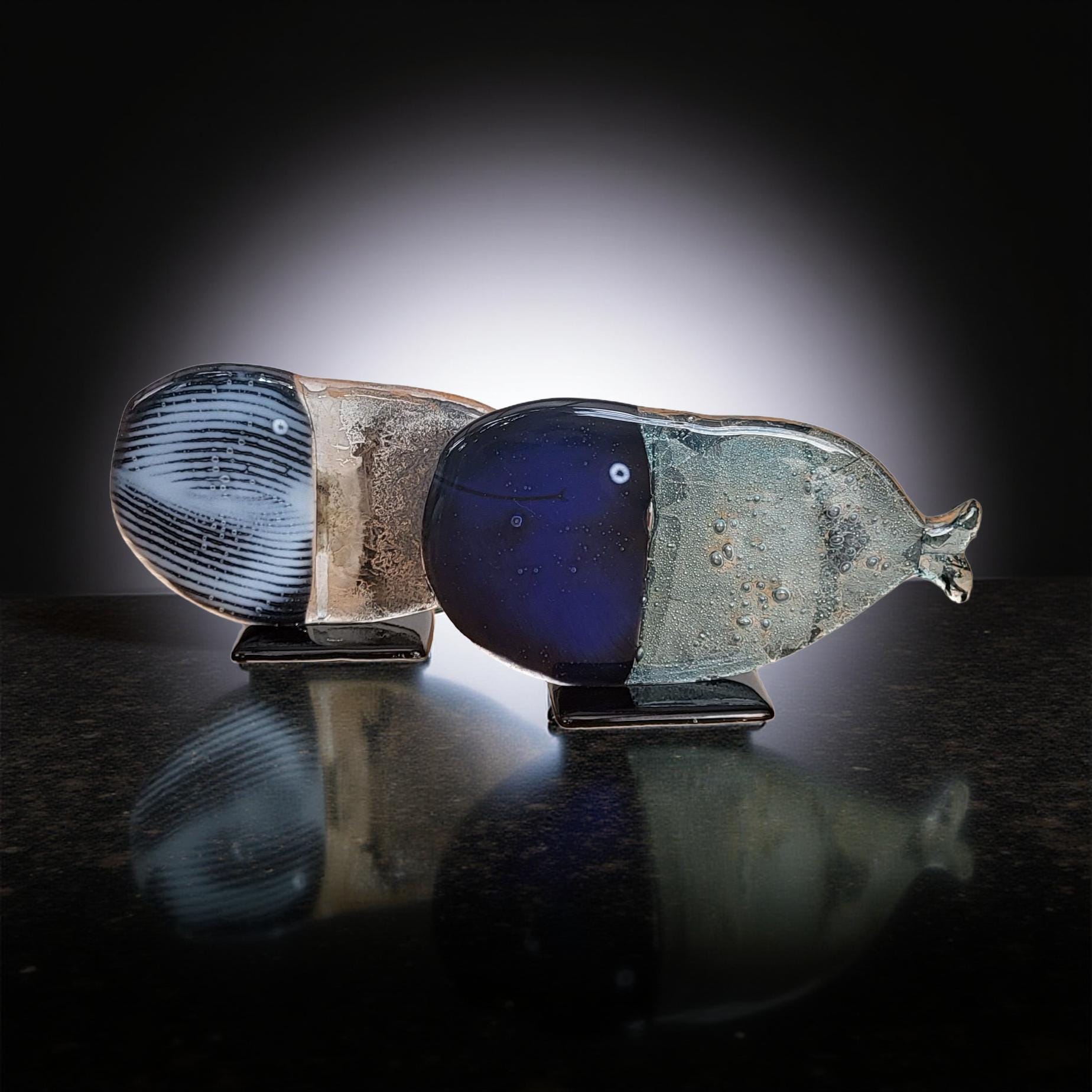 Two glass sculptures of fish, one blue and one gray, are displayed on a reflective surface against a dark background.