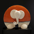 A ceramic elephant sculpture with a red and orange background and white trunk is displayed on a black stand against a black background.