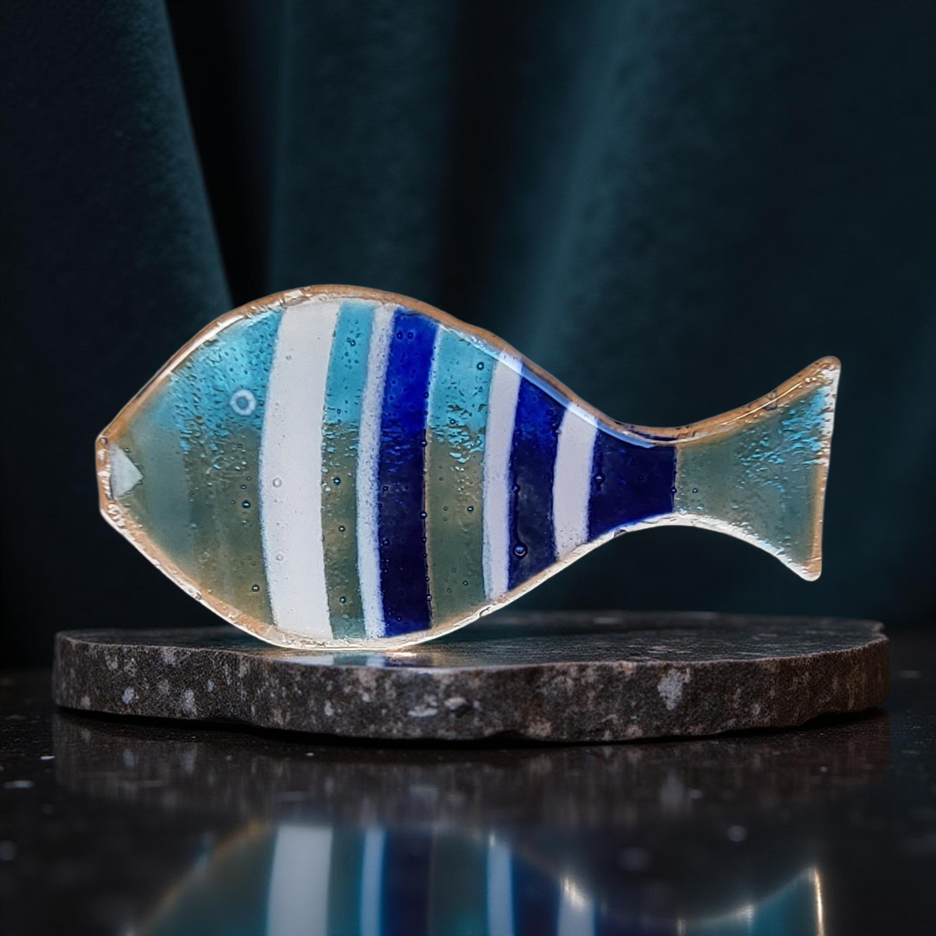 A colorful glass fish sculpture with blue and white stripes is displayed on a dark surface, possibly a stone or a black platform.