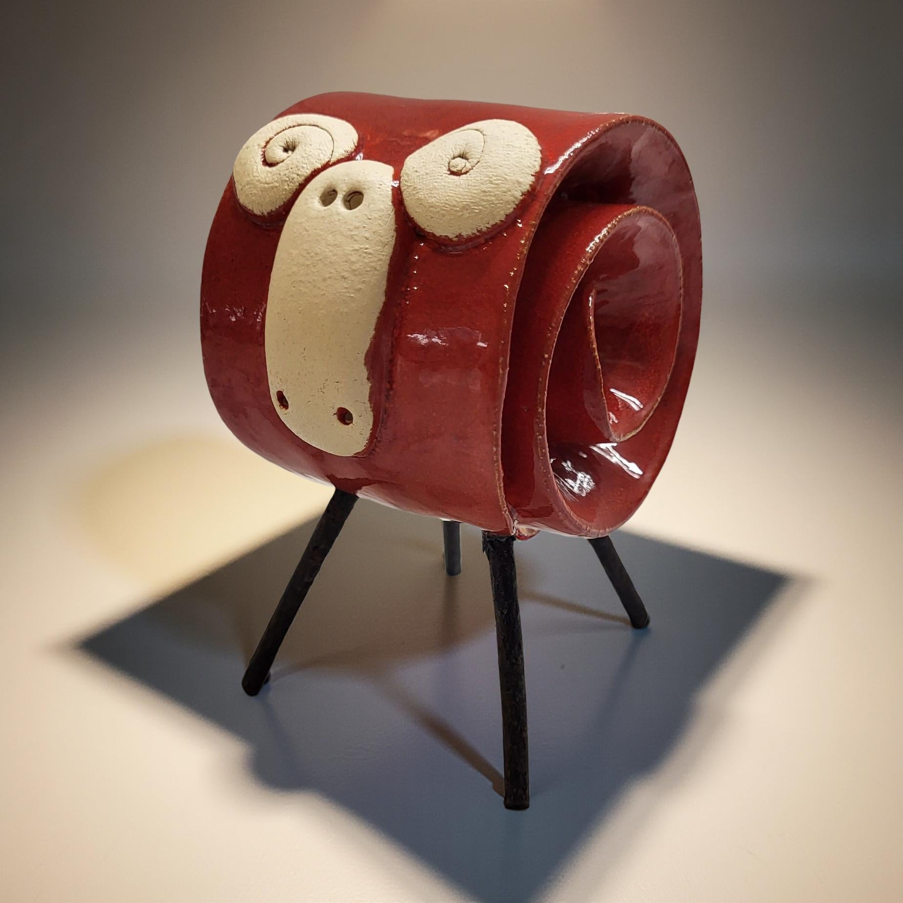 A red ceramic sculpture resembling a face with two white eyes and a small nose is displayed on a black metal stand against a plain background.