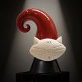 A ceramic cat sculpture with a red, curved finial and a white face, sitting on a black pedestal against a dark background.