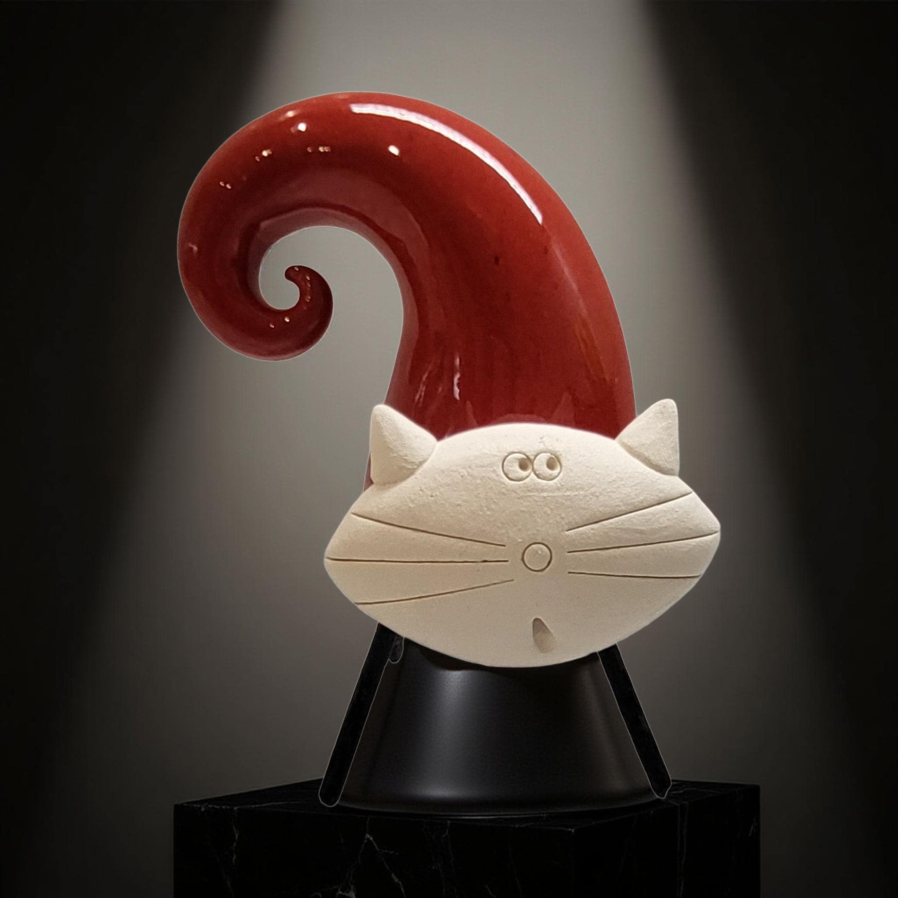 A ceramic cat sculpture with a red, curved finial and a white face, sitting on a black pedestal against a dark background.