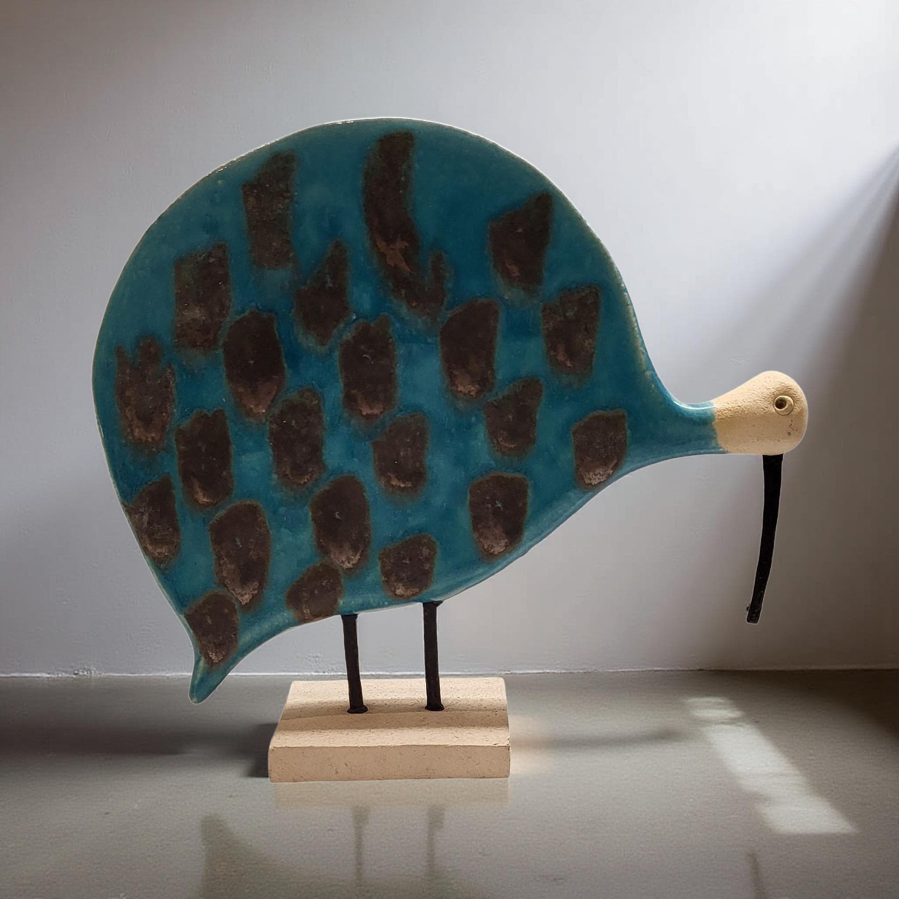 A blue ceramic sculpture of a bird-like creature with a brown patterned body and a beige beak, displayed on a wooden base.