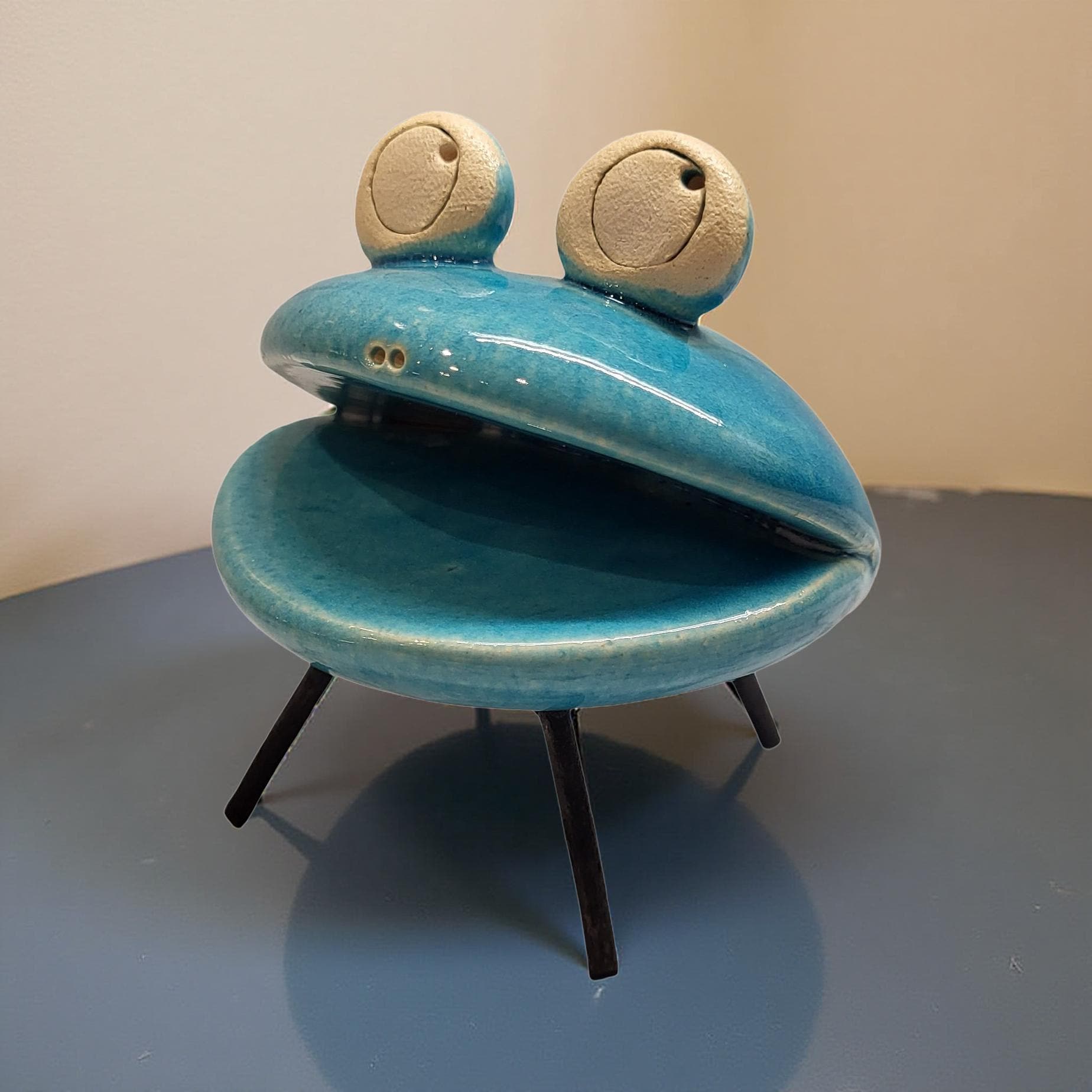 A blue ceramic frog figurine with large eyes and legs, sitting on a small stand.