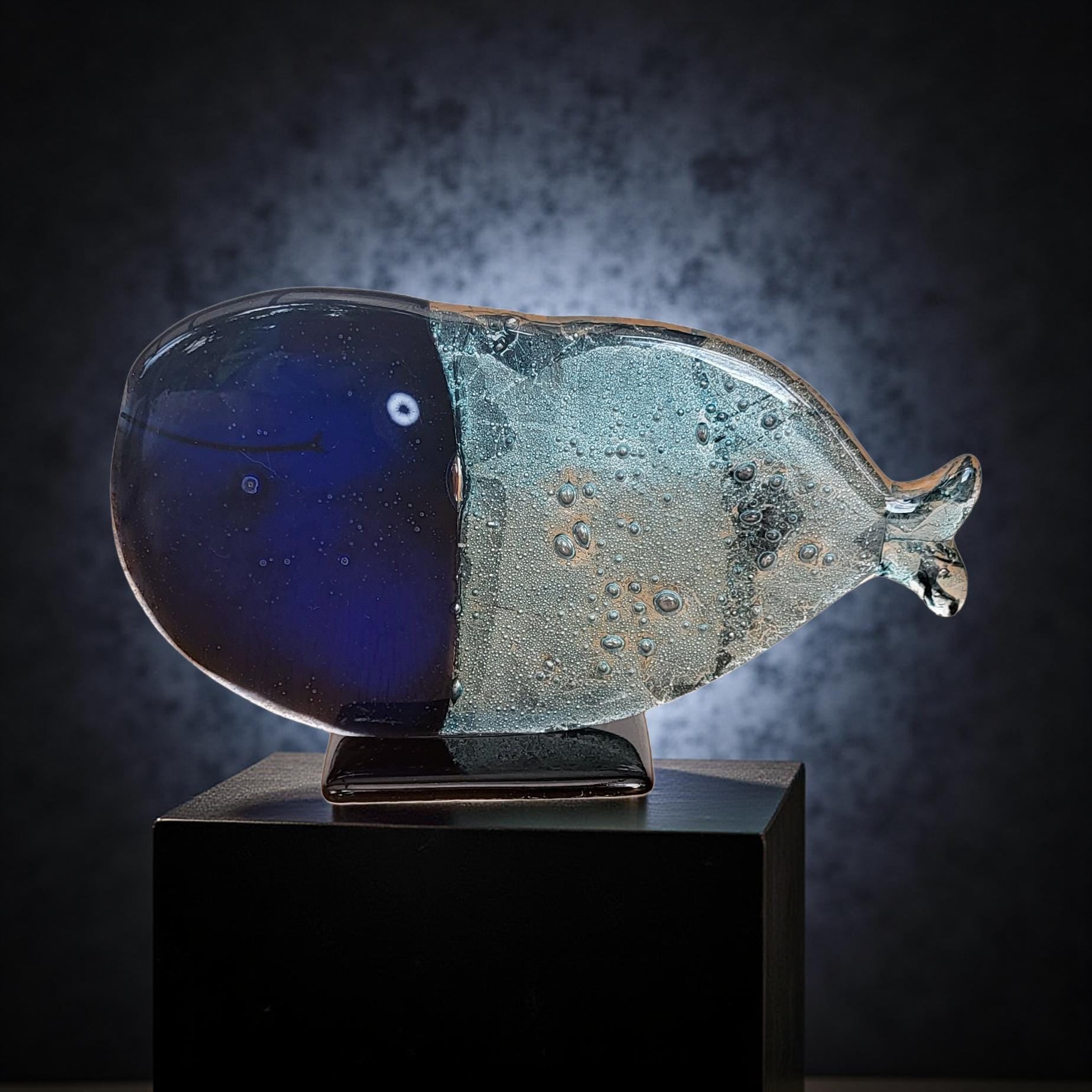 A glass sculpture of a whale, with a blue body and a white belly, is displayed on a black pedestal against a dark background.