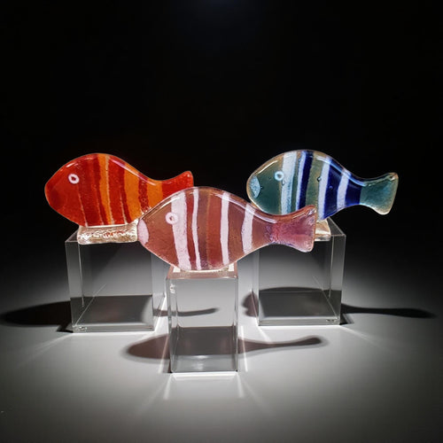 Three colorful glass fish figurines are displayed on glass stands against a dark background.