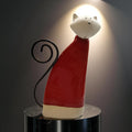 A ceramic cat figurine wearing a red sweater and standing on a black pedestal.