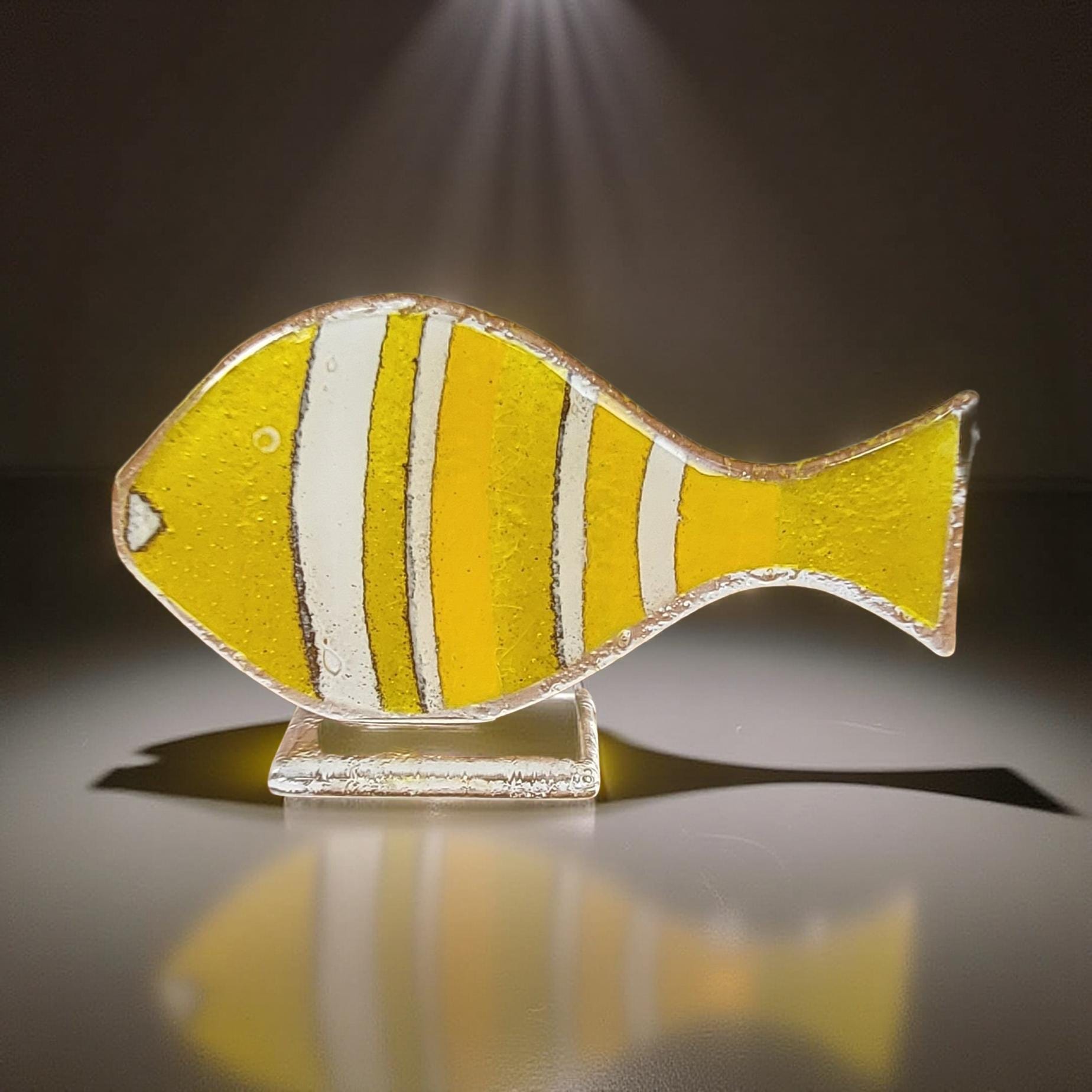 A yellow and white striped fish sculpture is displayed on a reflective surface, casting a shadow on the ground.