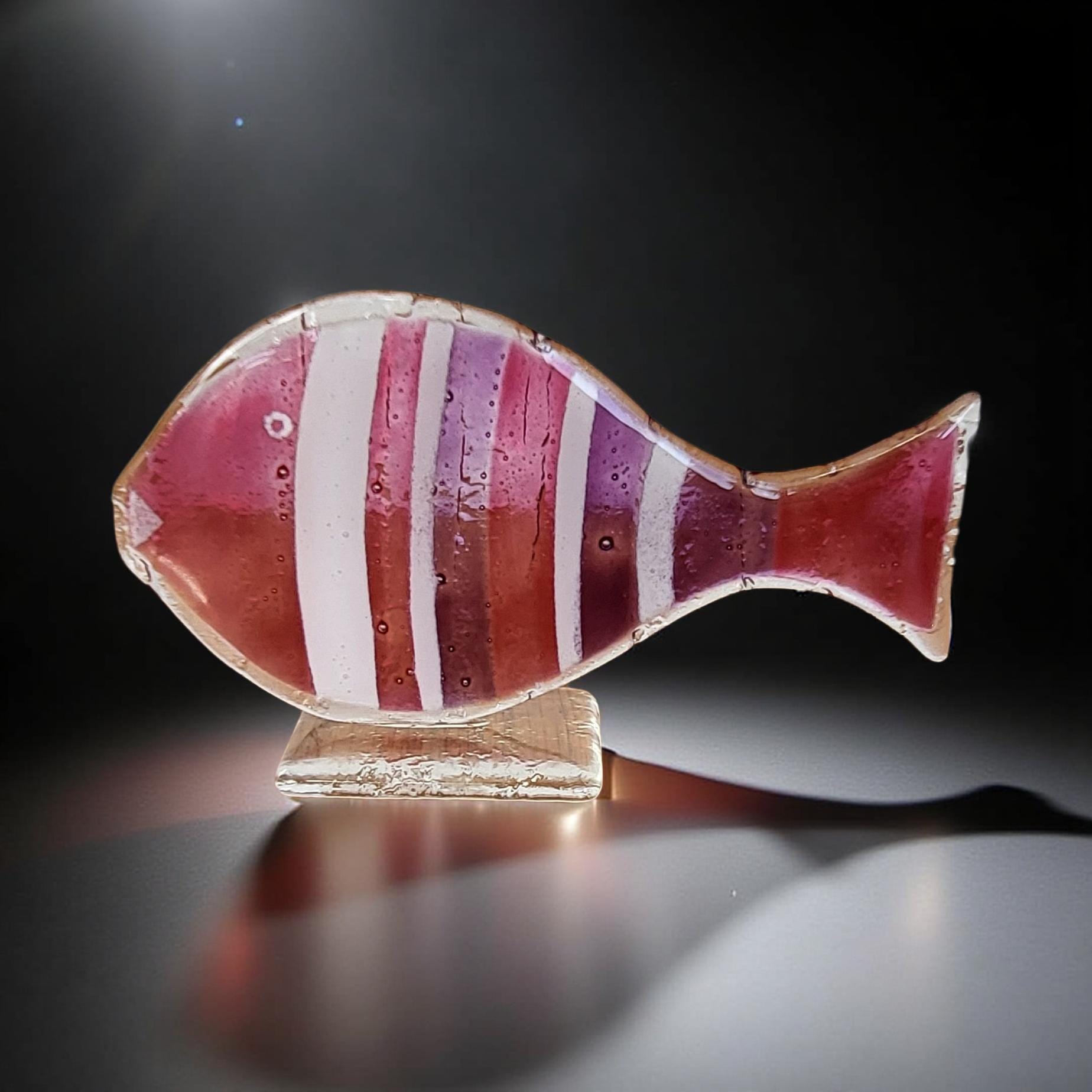 A colorful glass fish sculpture with red and white stripes is displayed on a stand against a dark background.