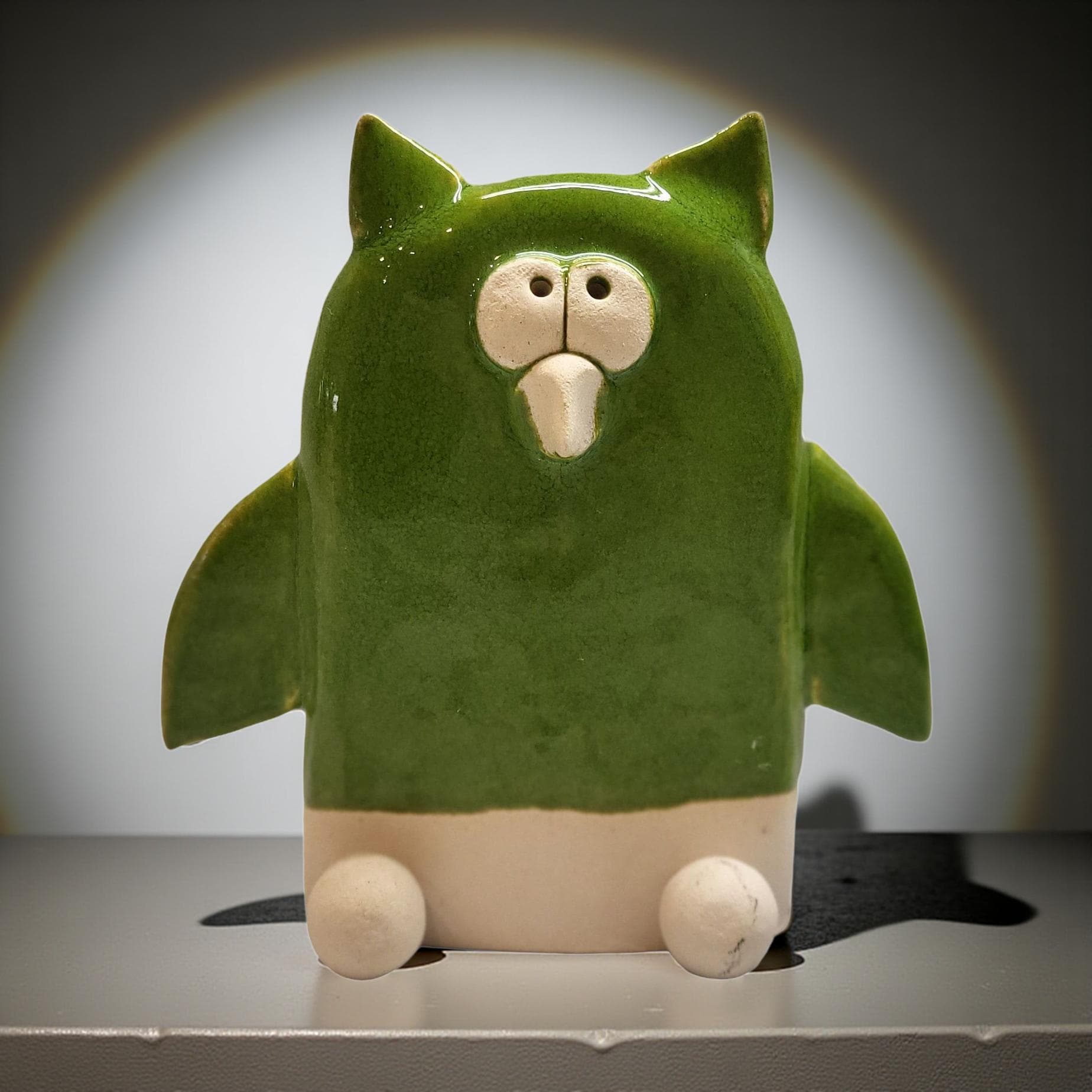 A green ceramic owl figurine with a white face and beak is sitting on a white surface against a white background.