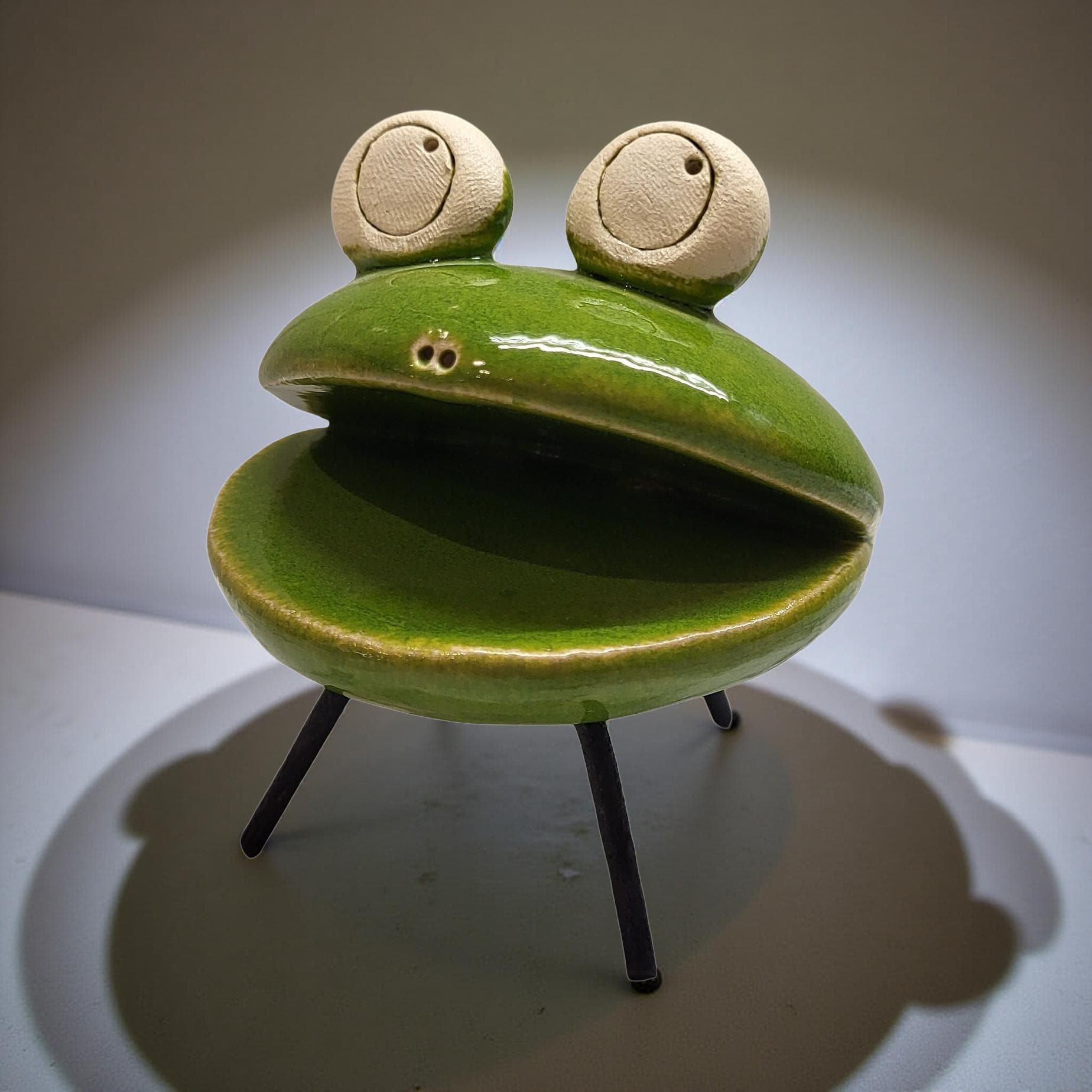 A green ceramic frog figurine with large eyes and a small mouth is sitting on a white surface.