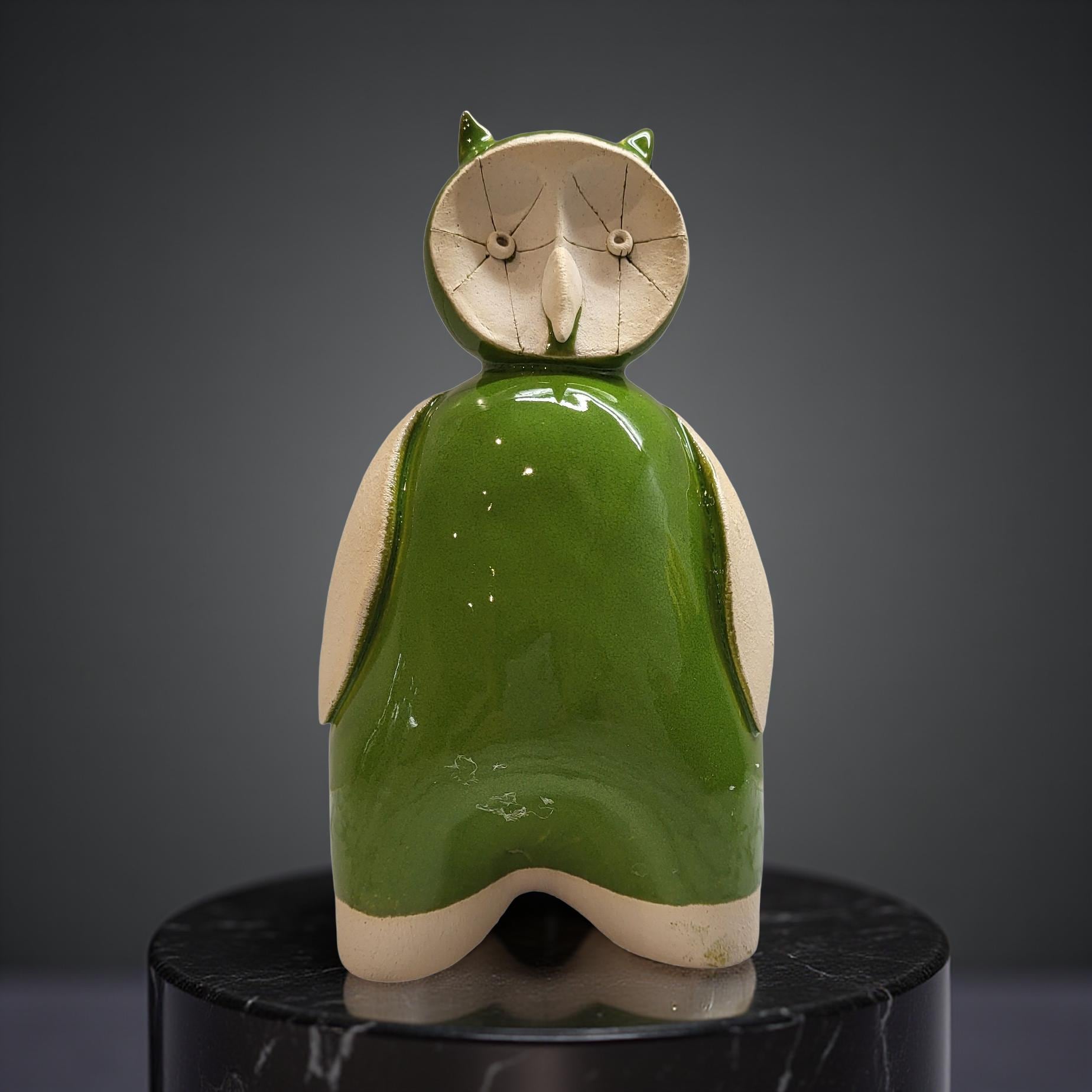 A green ceramic owl figurine with a white face and beak is sitting on a black pedestal against a gray background.