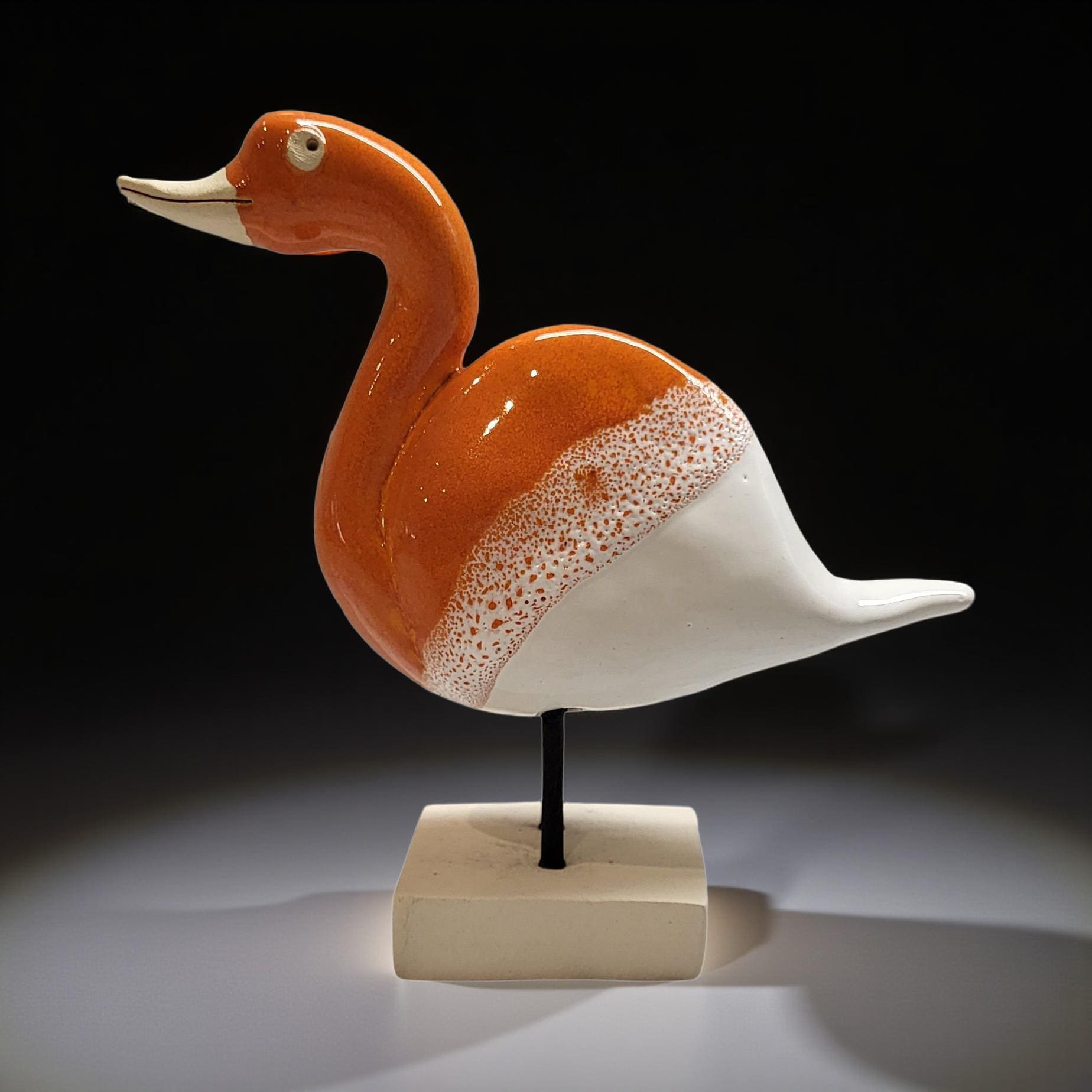 A ceramic sculpture of a duck, featuring an orange body with white speckles, is displayed on a small square base against a dark background.
