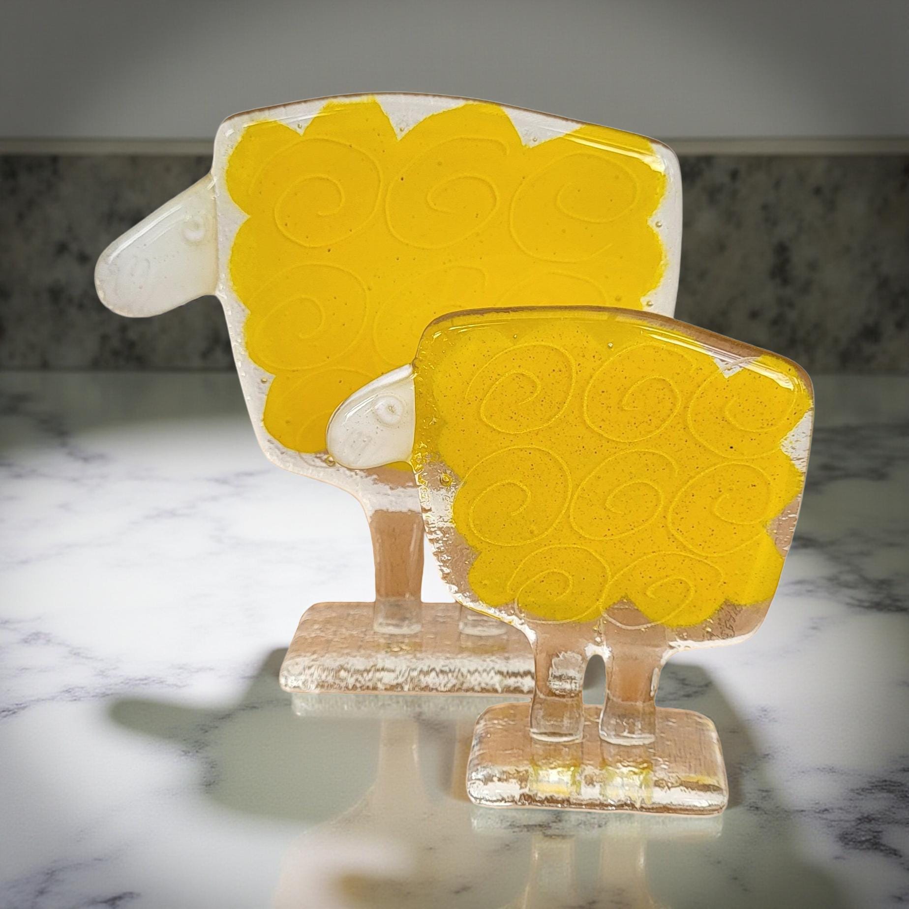 Two glass sculptures of sheep, one larger and one smaller, both with yellow and white colors and spiral designs on their bodies.