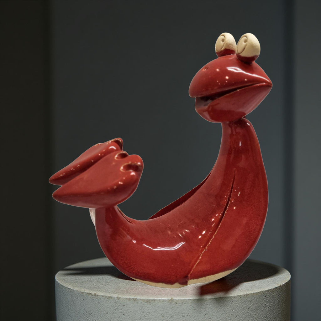 A red ceramic sculpture of a smiling fish with large eyes and a long, curved body is displayed on a gray pedestal.