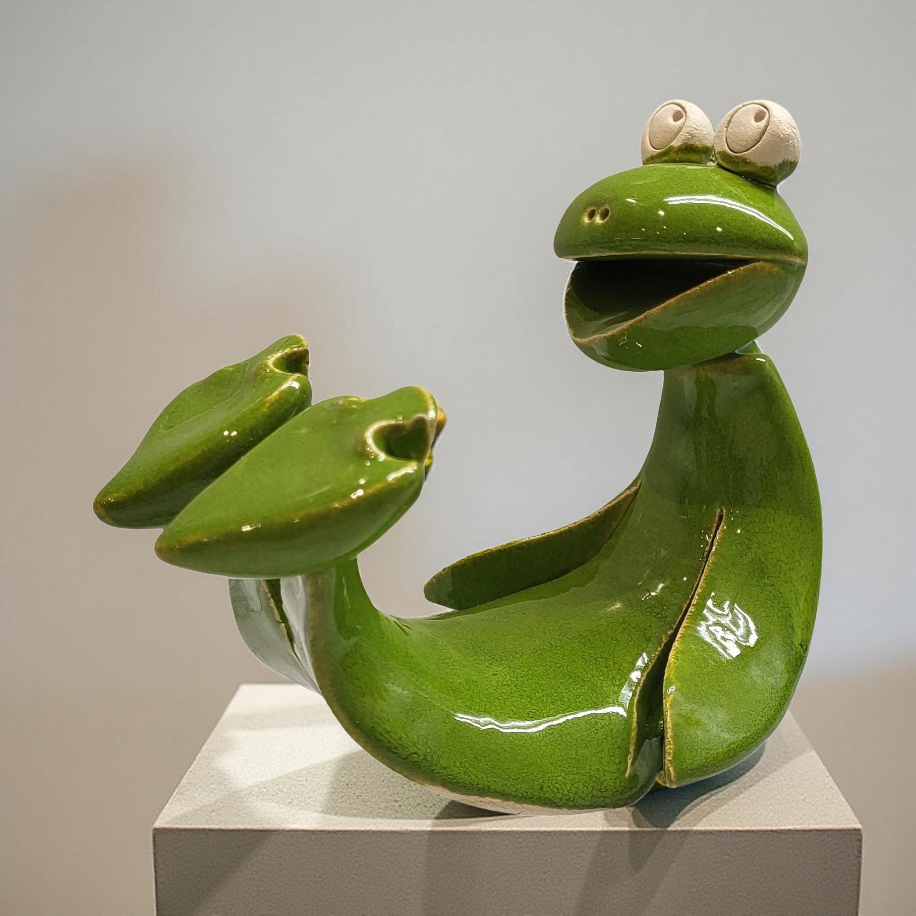 A green ceramic frog sculpture with large eyes and a wide smile, sitting on a white pedestal.