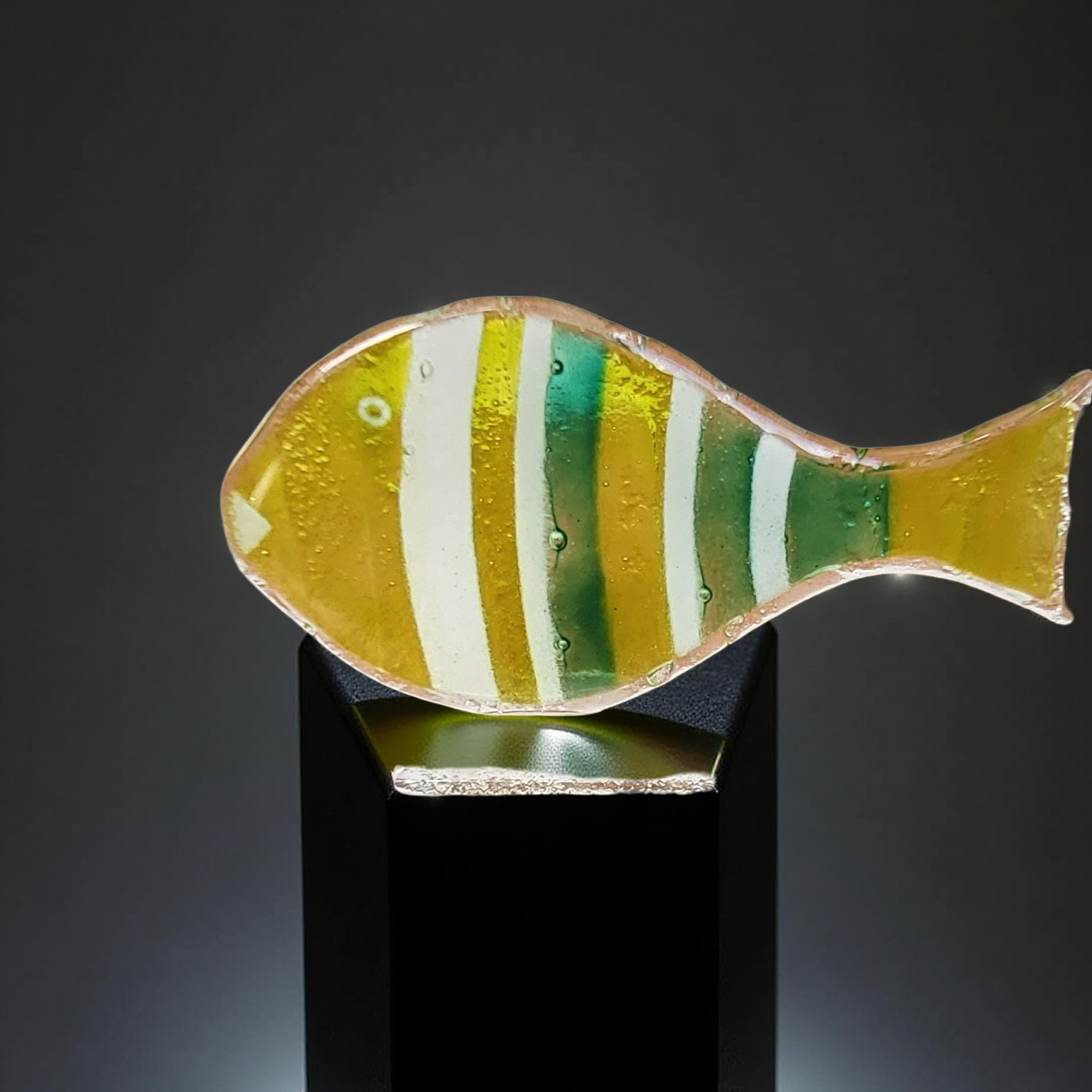 A colorful glass fish sculpture is displayed on a black pedestal against a dark background.