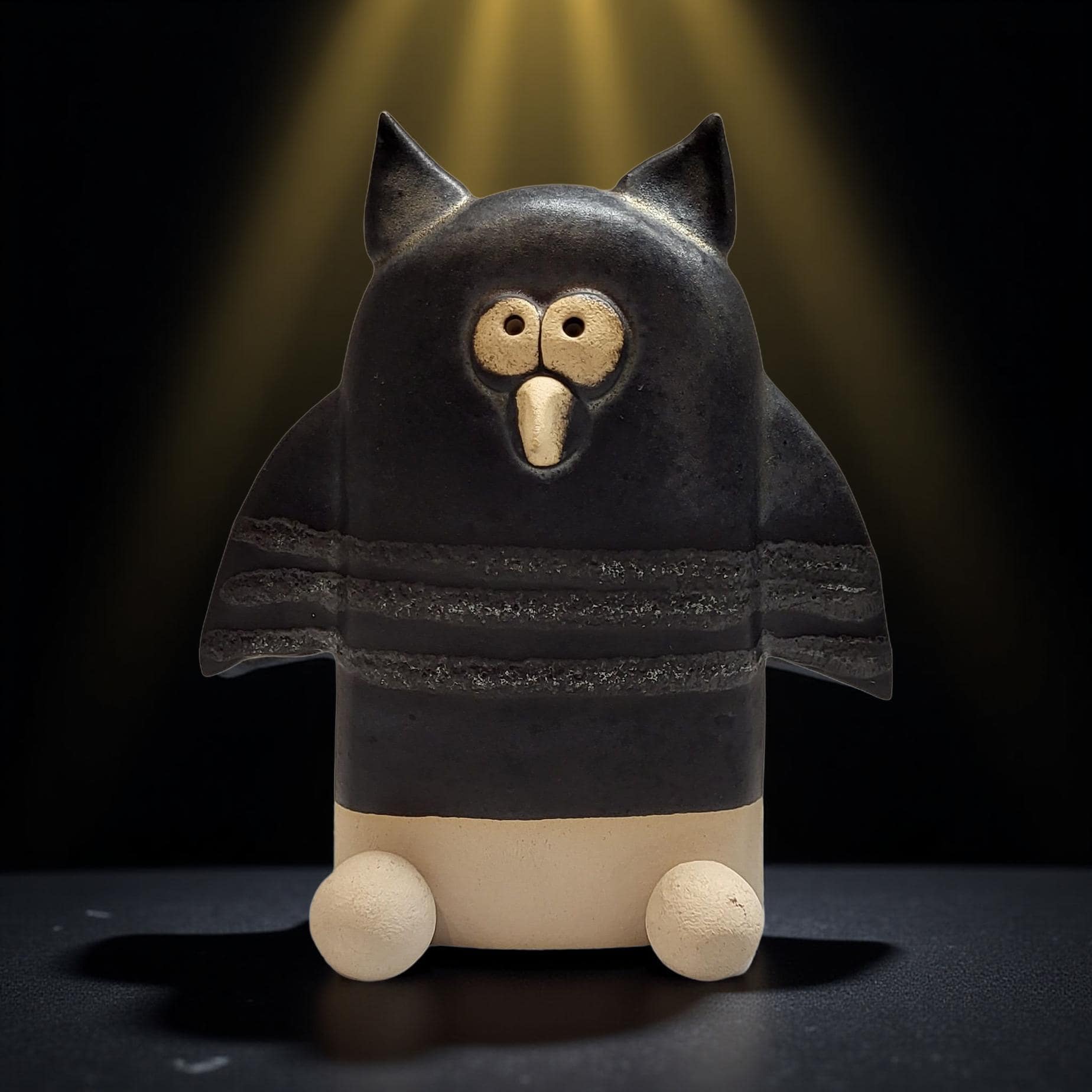A ceramic owl figurine with a black body and white accents, sitting on a dark surface and illuminated by a spotlight.