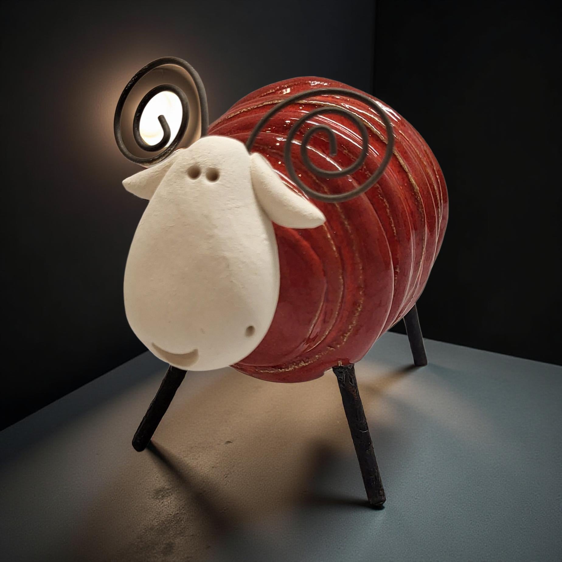 A red ceramic sheep figurine with a white face and black legs is displayed on a gray surface, illuminated by a light source.