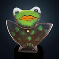 A glass frog sculpture with a green body and orange tongue, wearing a crown and standing on a black stand against a dark background.