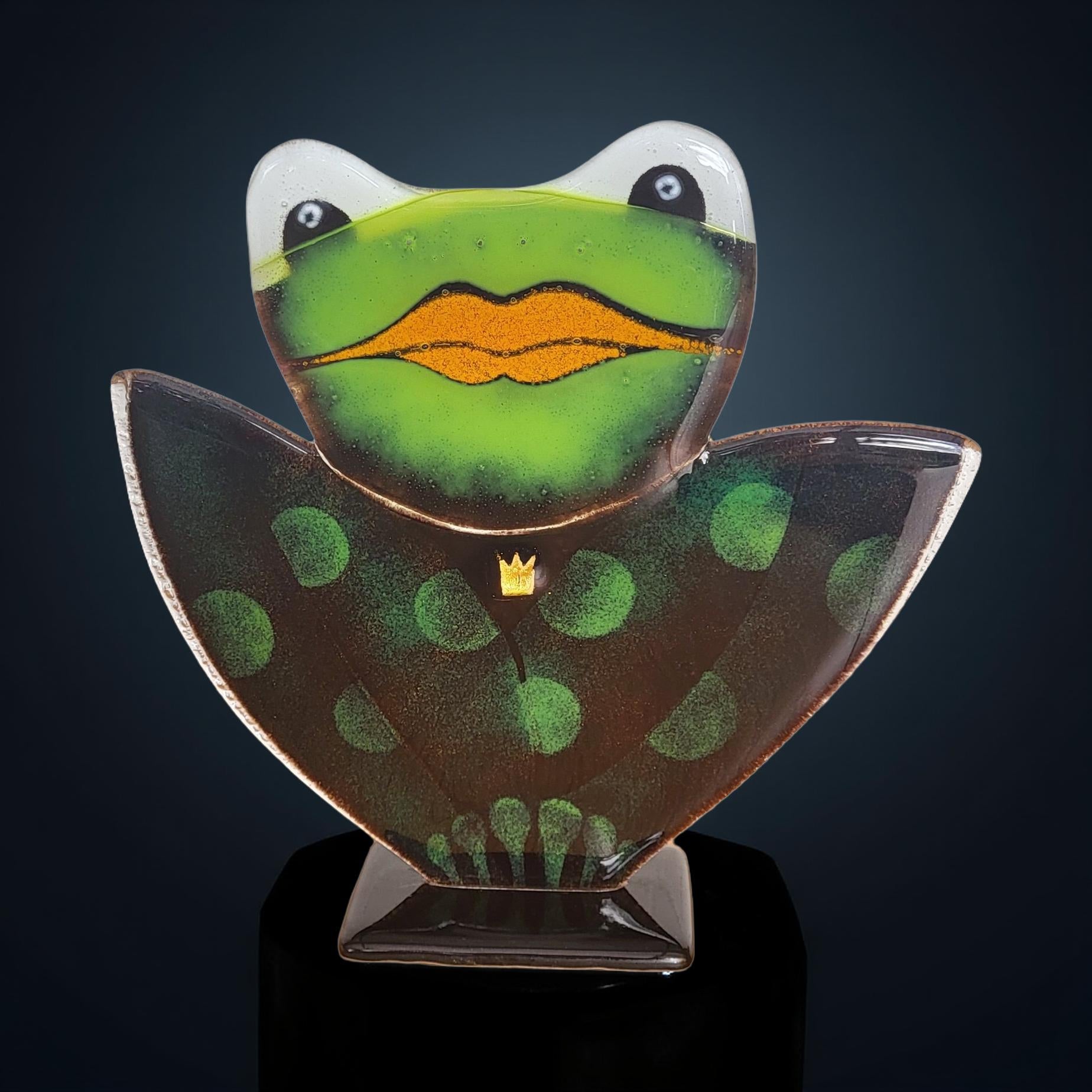 A glass frog sculpture with a green body and orange tongue, wearing a crown and standing on a black stand against a dark background.